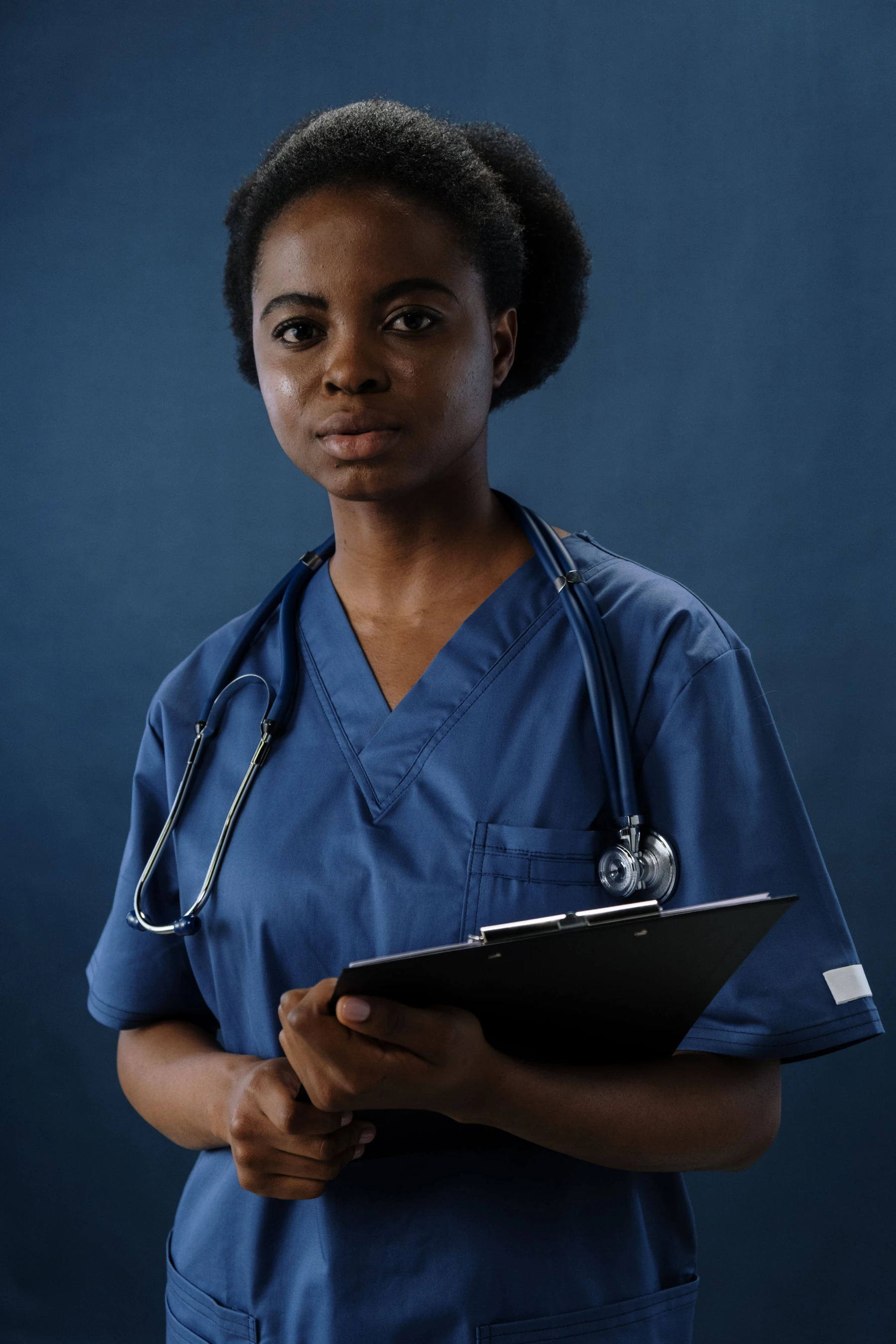 Medical assistant in navy blue scrubs with a stethoscope holding a clipboard against a blue background