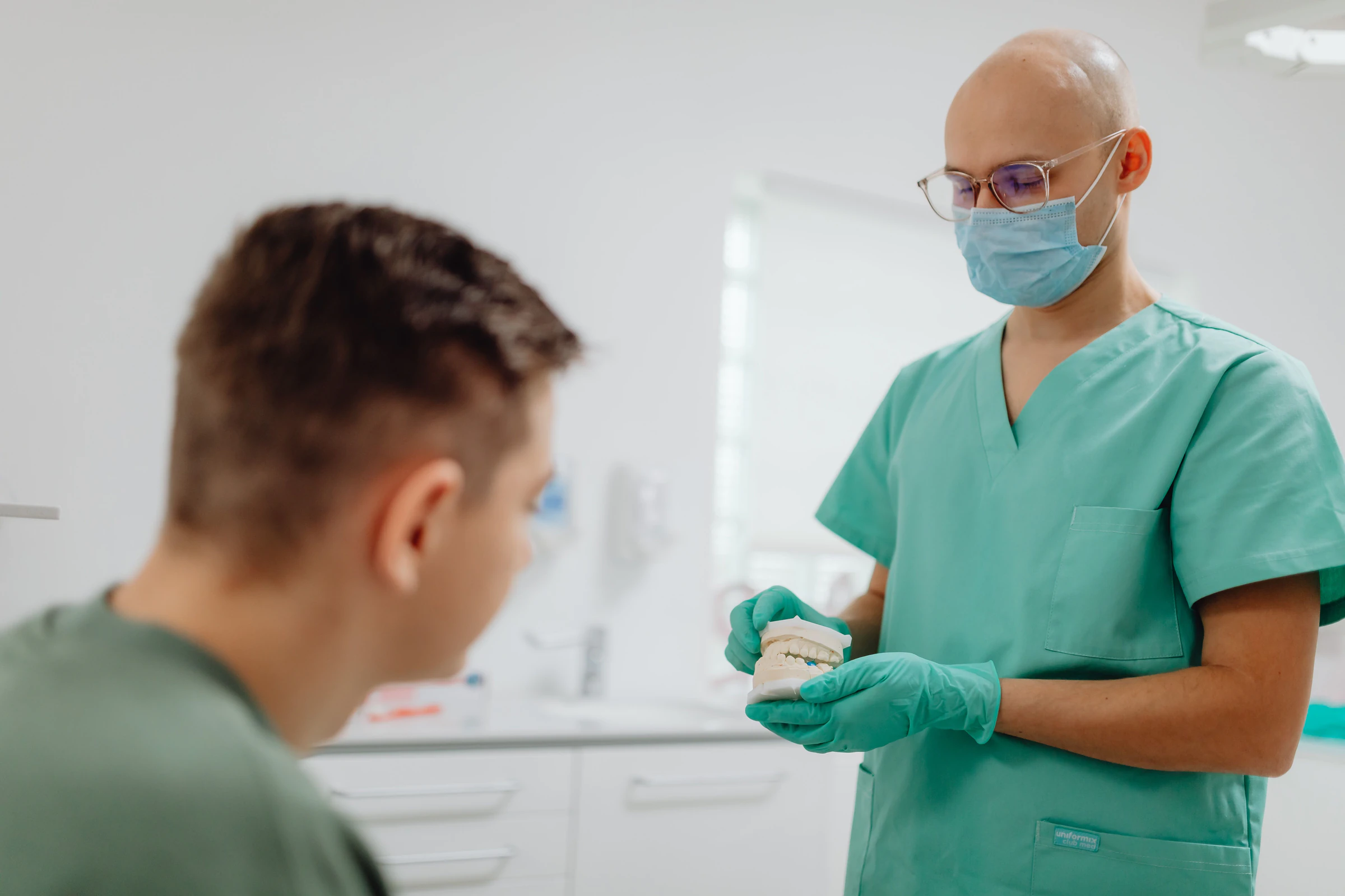 Dentist in green scrubs, mask and gloves explaining a procedure to a young patient in a clinic