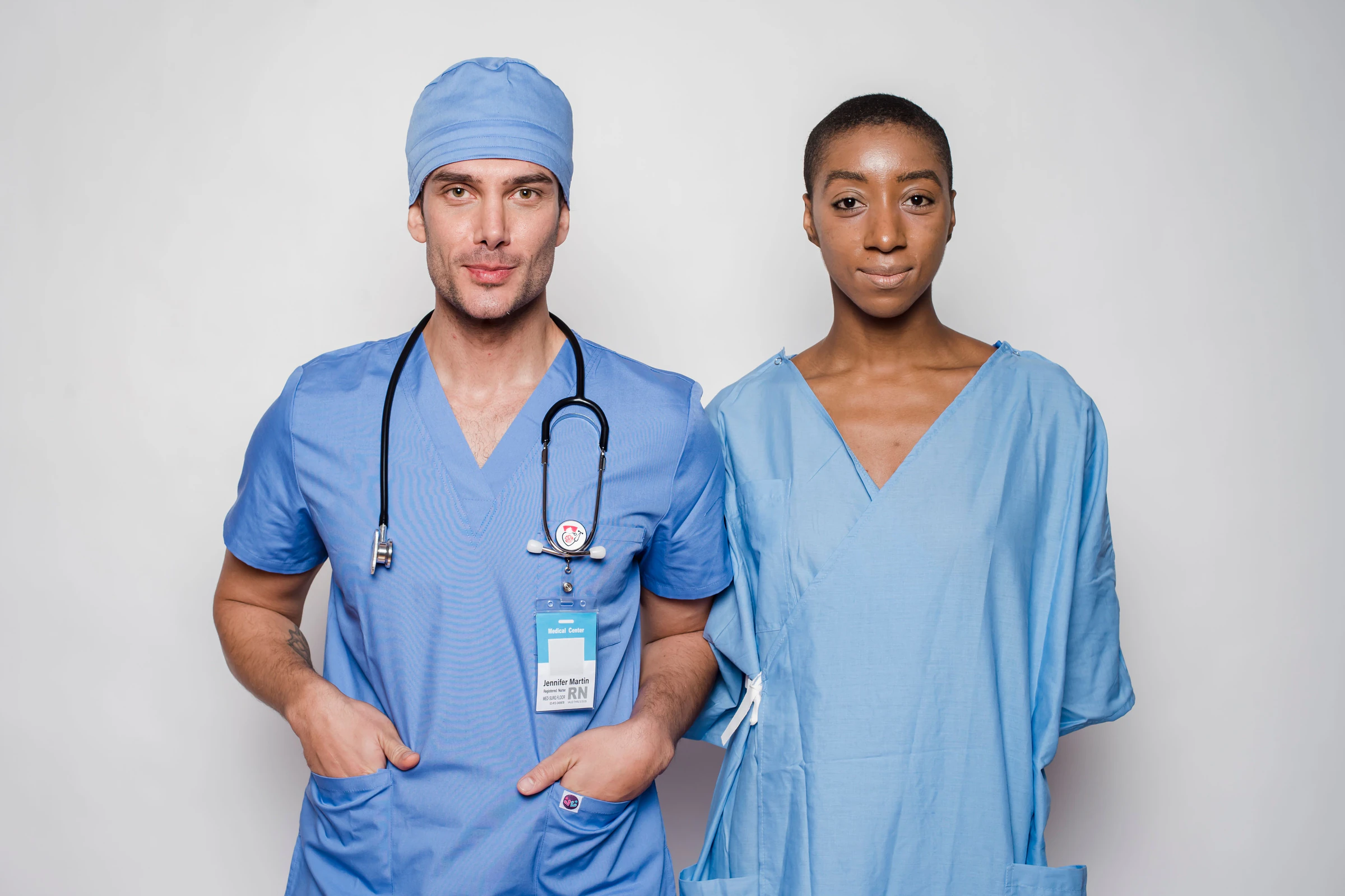 Male doctor in blue scrubs, scrub cap and stethoscope standing beside a female colleague in light blue scrubs