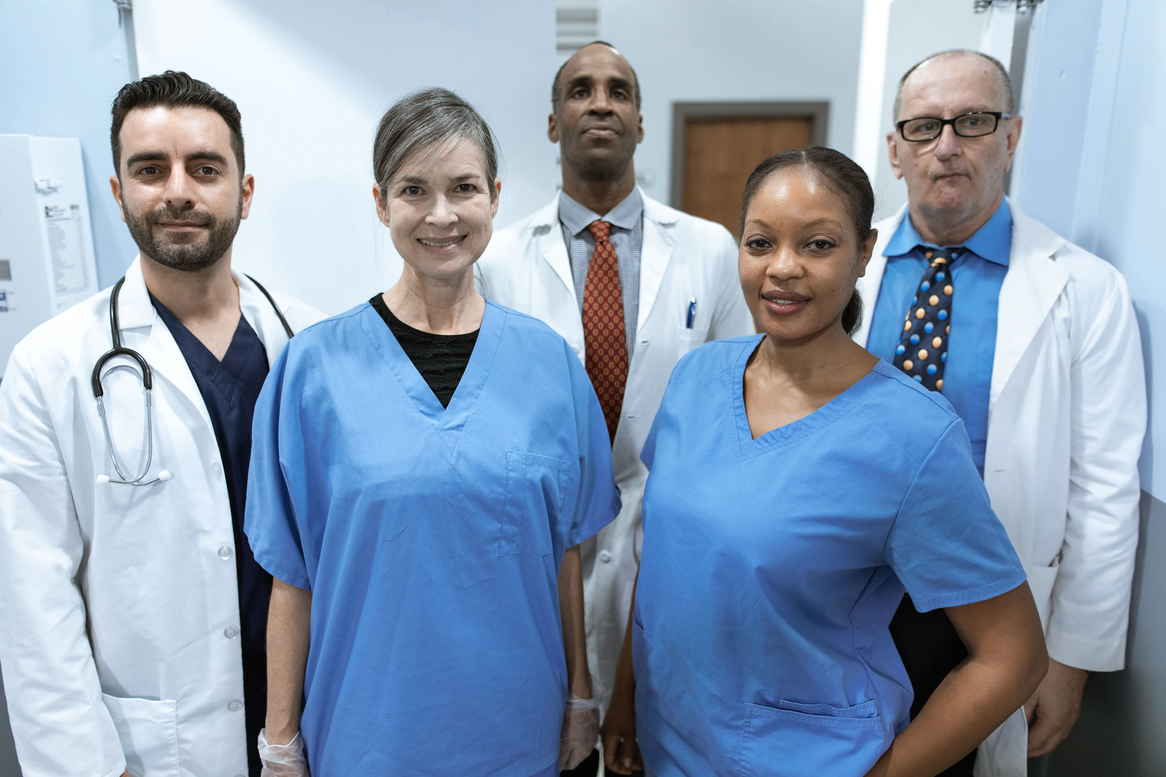 Group of smiling healthcare workers in blue scrubs and white lab coats posing in a hospital corridor
