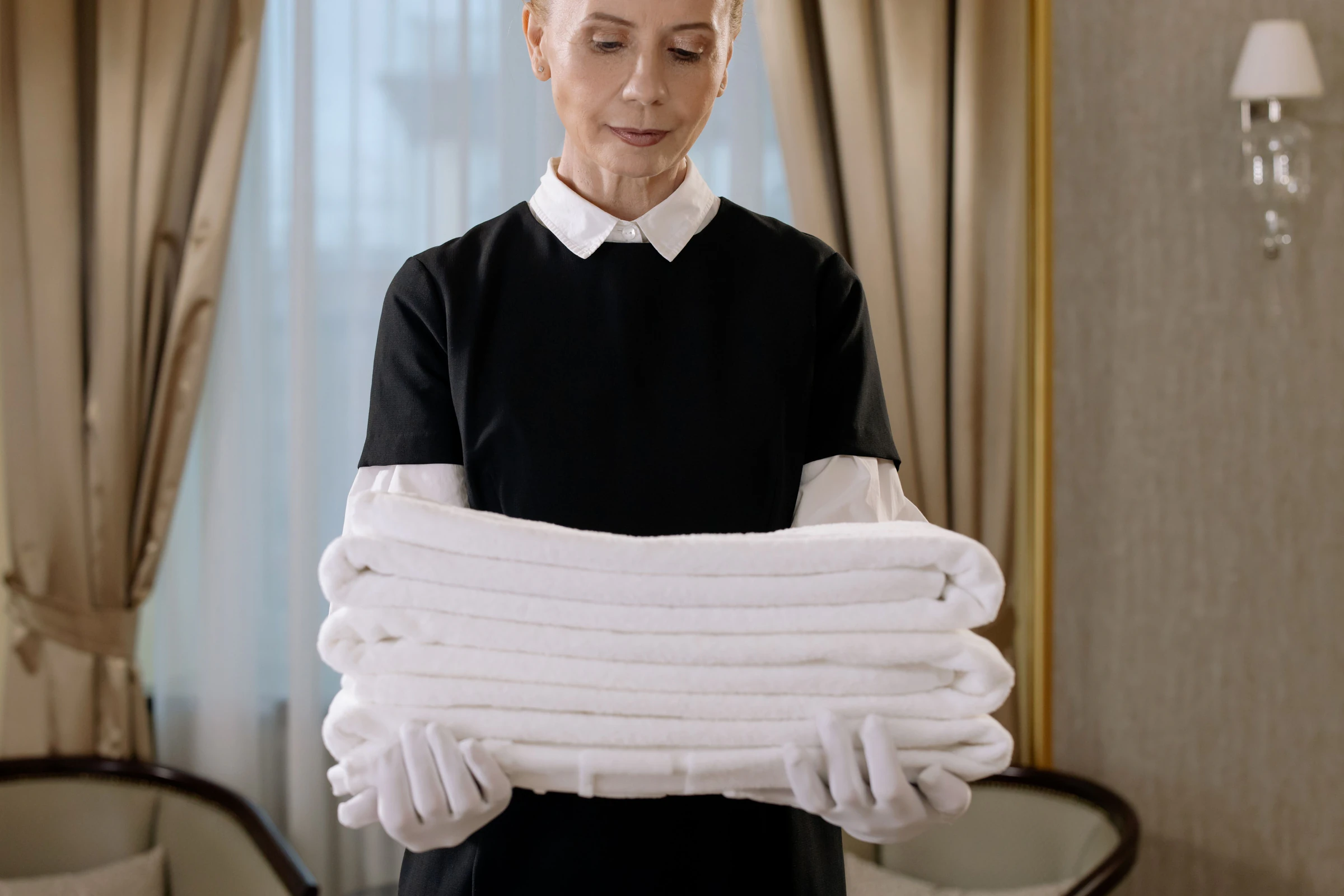 Hotel housekeeper in a black uniform with white collar and gloves holding a stack of fresh towels