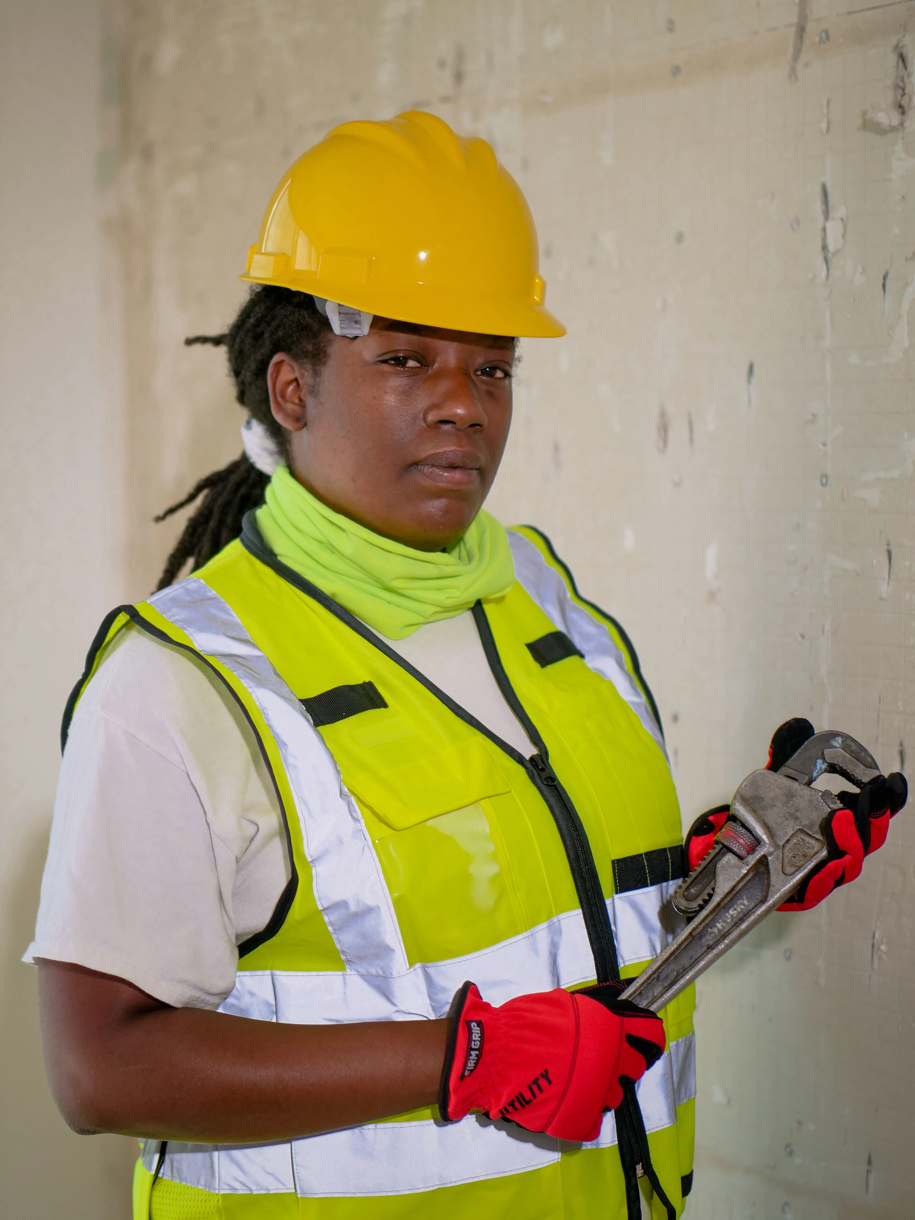 Tradeswoman in a yellow hard hat and hi-vis vest holding a large pipe wrench indoors
