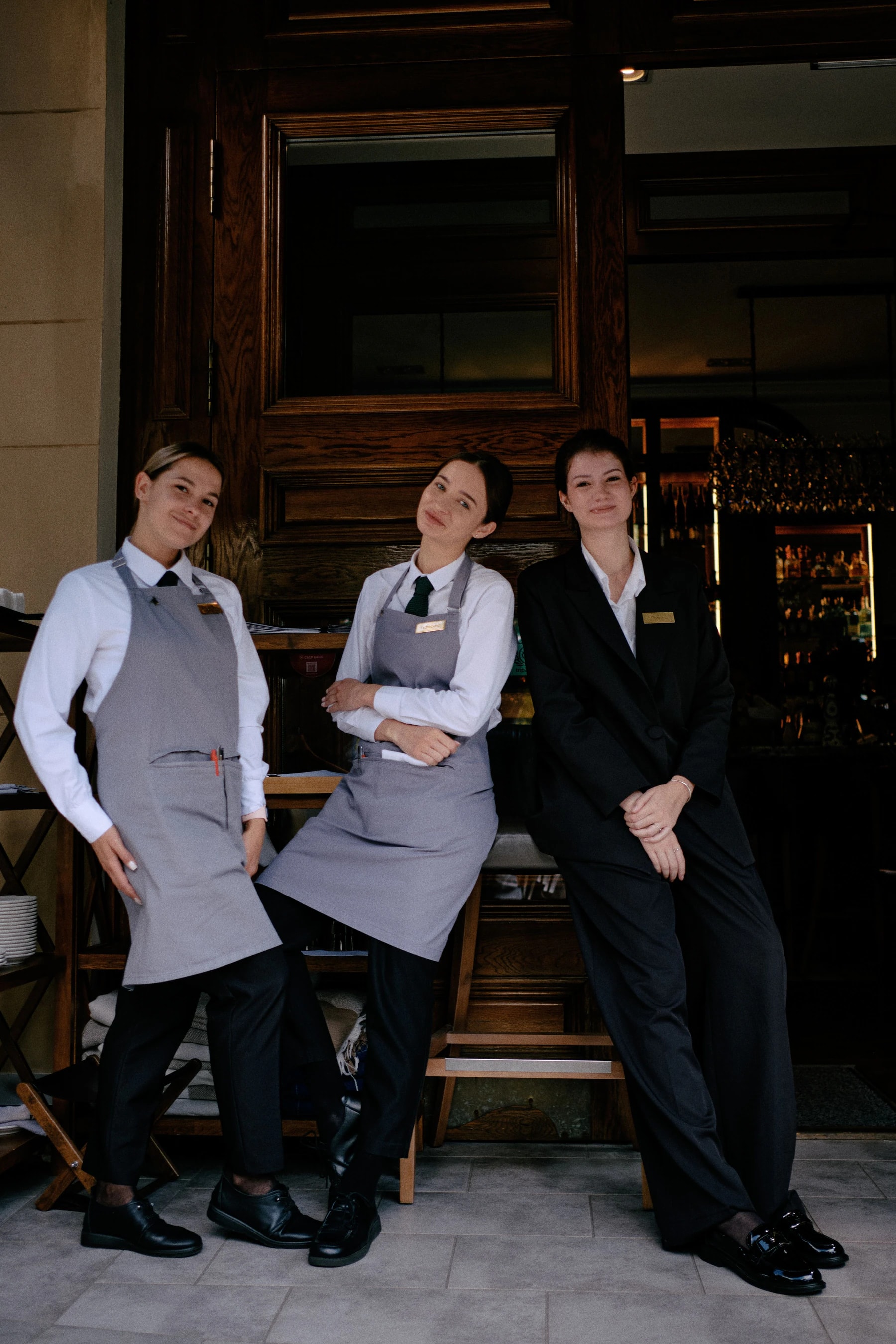 Three restaurant staff in white shirts, gray bib aprons and a black suit posing at a restaurant entrance