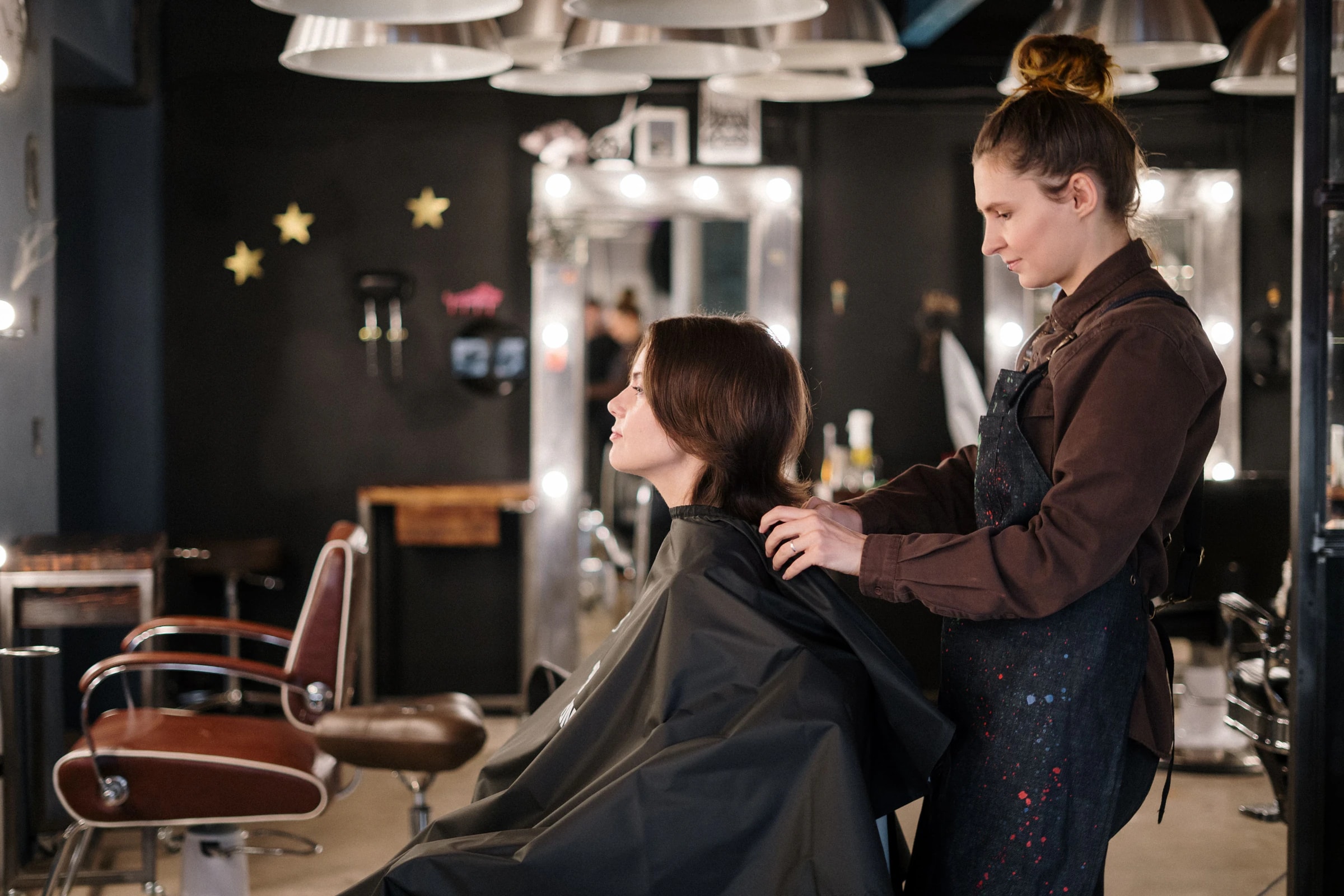 Hair stylist in a dark apron preparing a client draped in a cape at a modern salon station