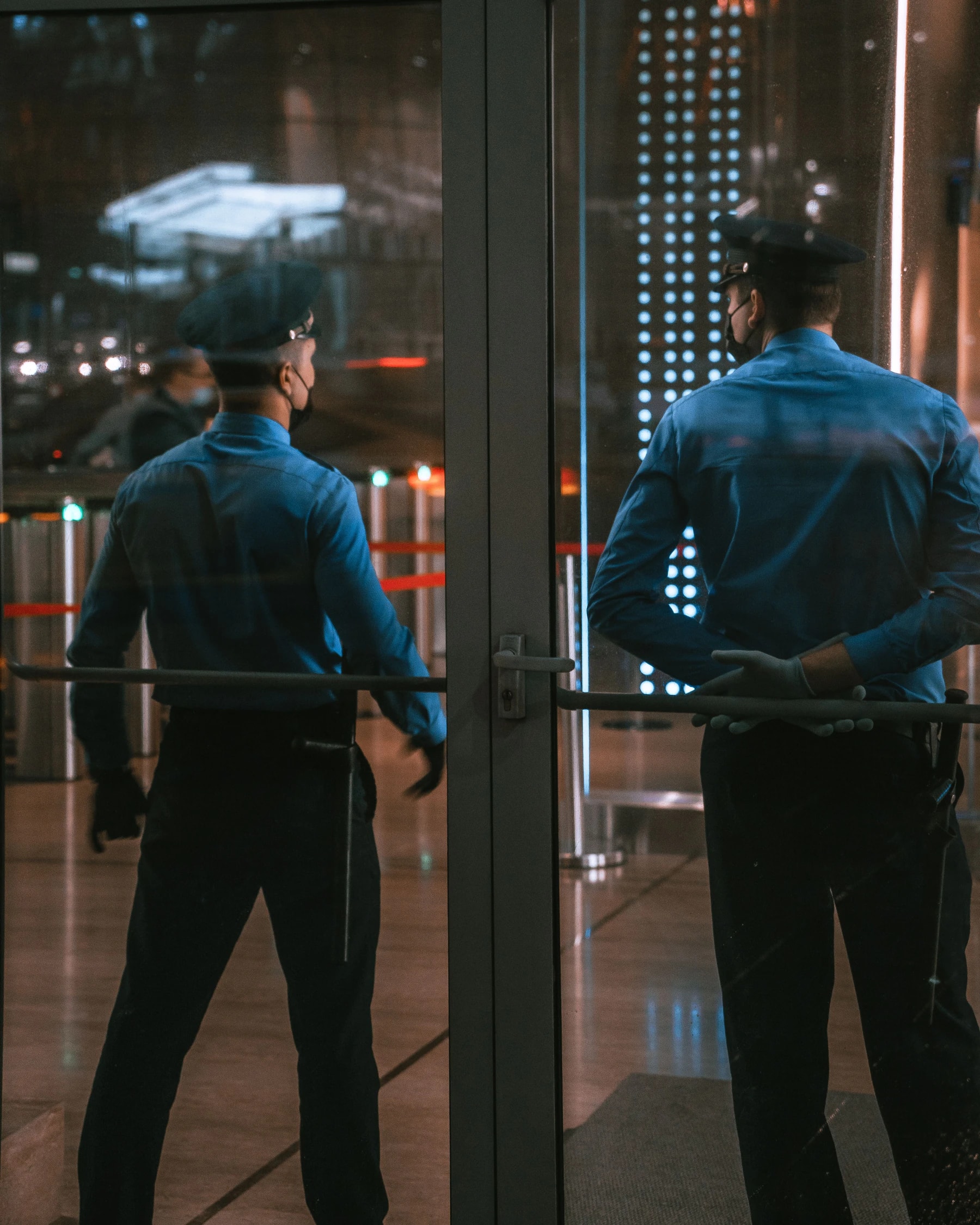 Two security guards in blue long-sleeve uniforms and caps standing watch at a building glass entrance