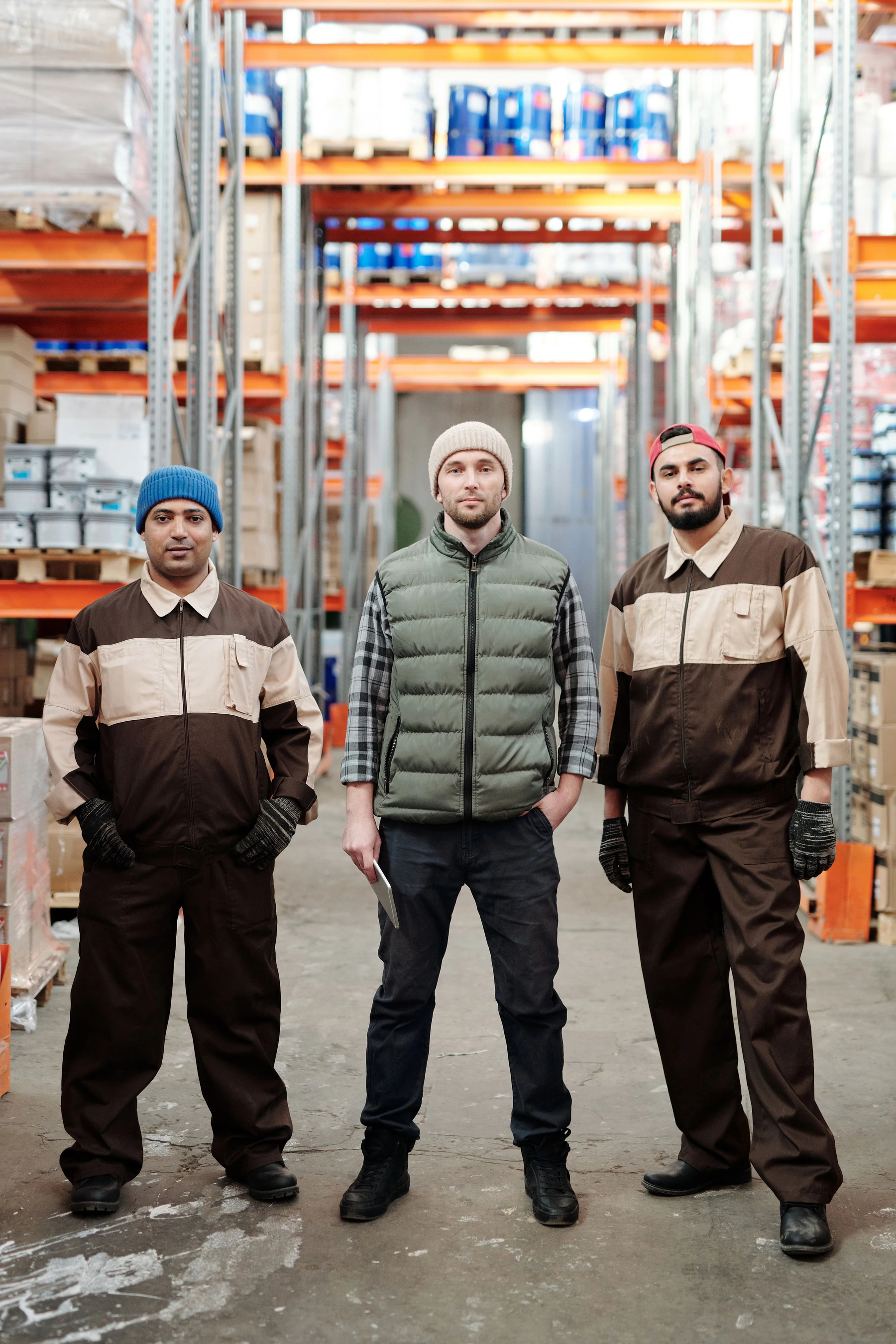 Three warehouse workers, two in brown and beige work uniforms, standing in an aisle of tall racks