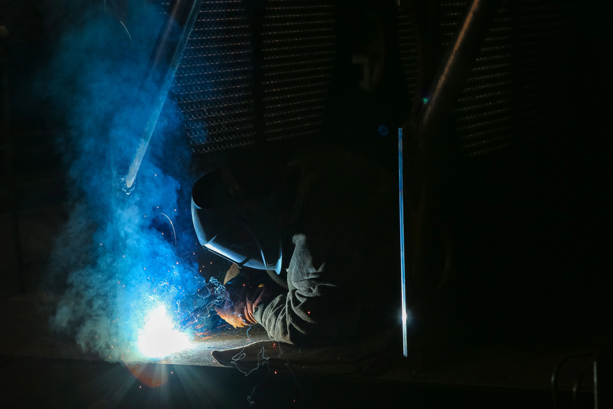 Welder in protective gloves and welding helmet working with a bright blue arc and sparks in a dark shop