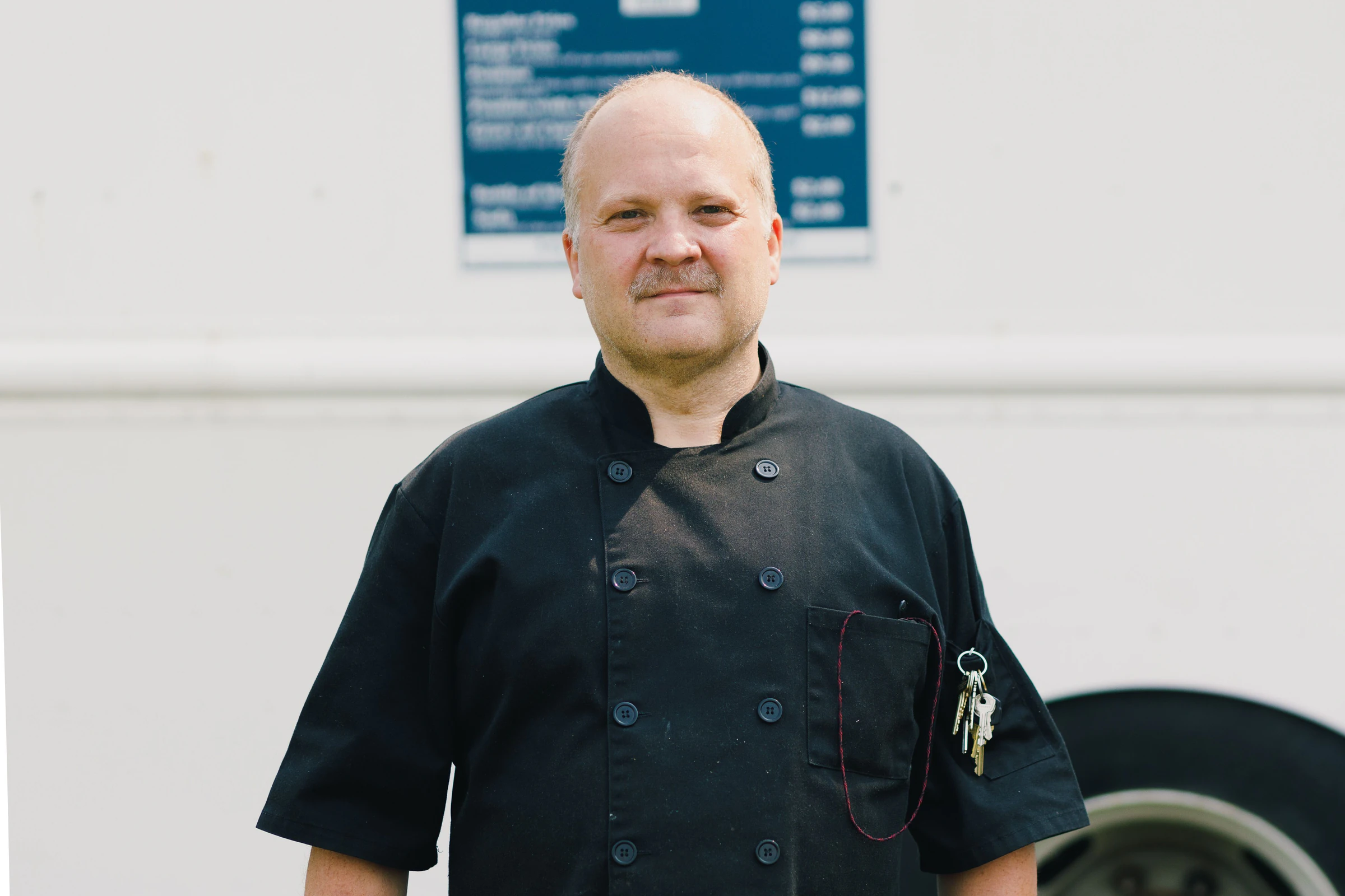 Cook in a black short-sleeve button-front chef shirt with a sleeve pocket standing outside a food truck