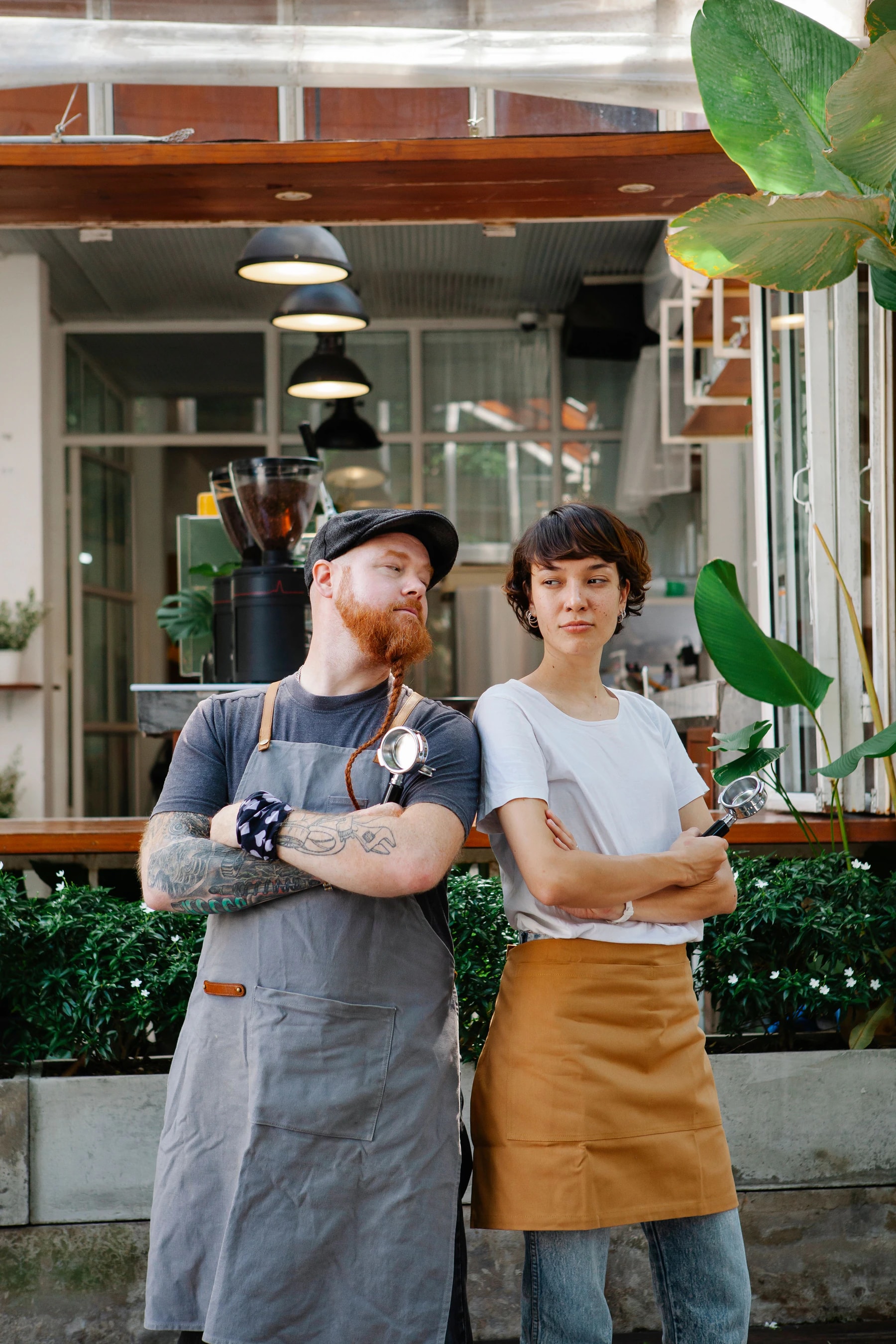 Two cafe workers in gray and tan canvas aprons standing with arms crossed outside a coffee shop