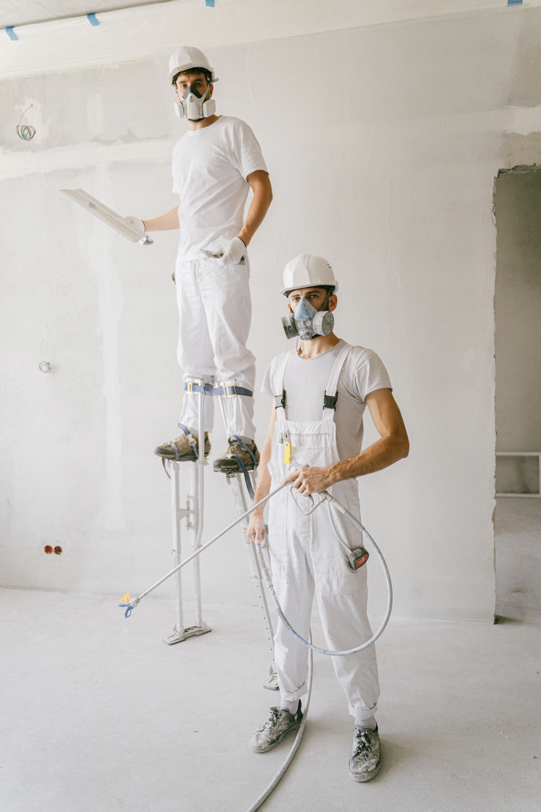 Two painters in white work pants, white bib overalls and respirators working in a white room