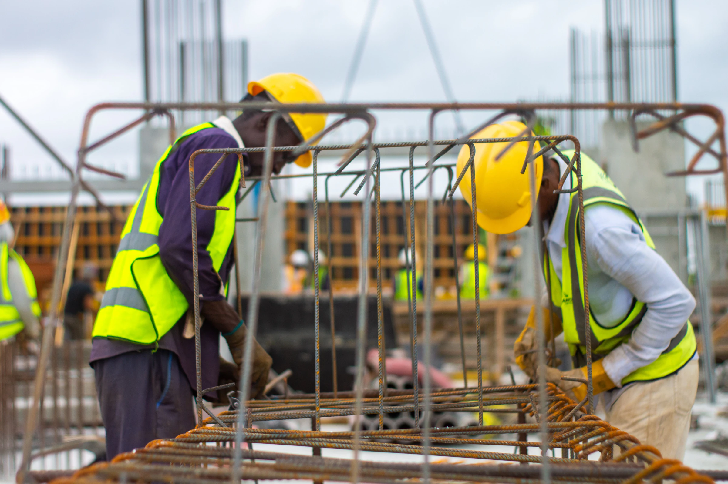 Two construction workers in yellow-green hi-vis safety vests and yellow hard hats tying rebar cages on site