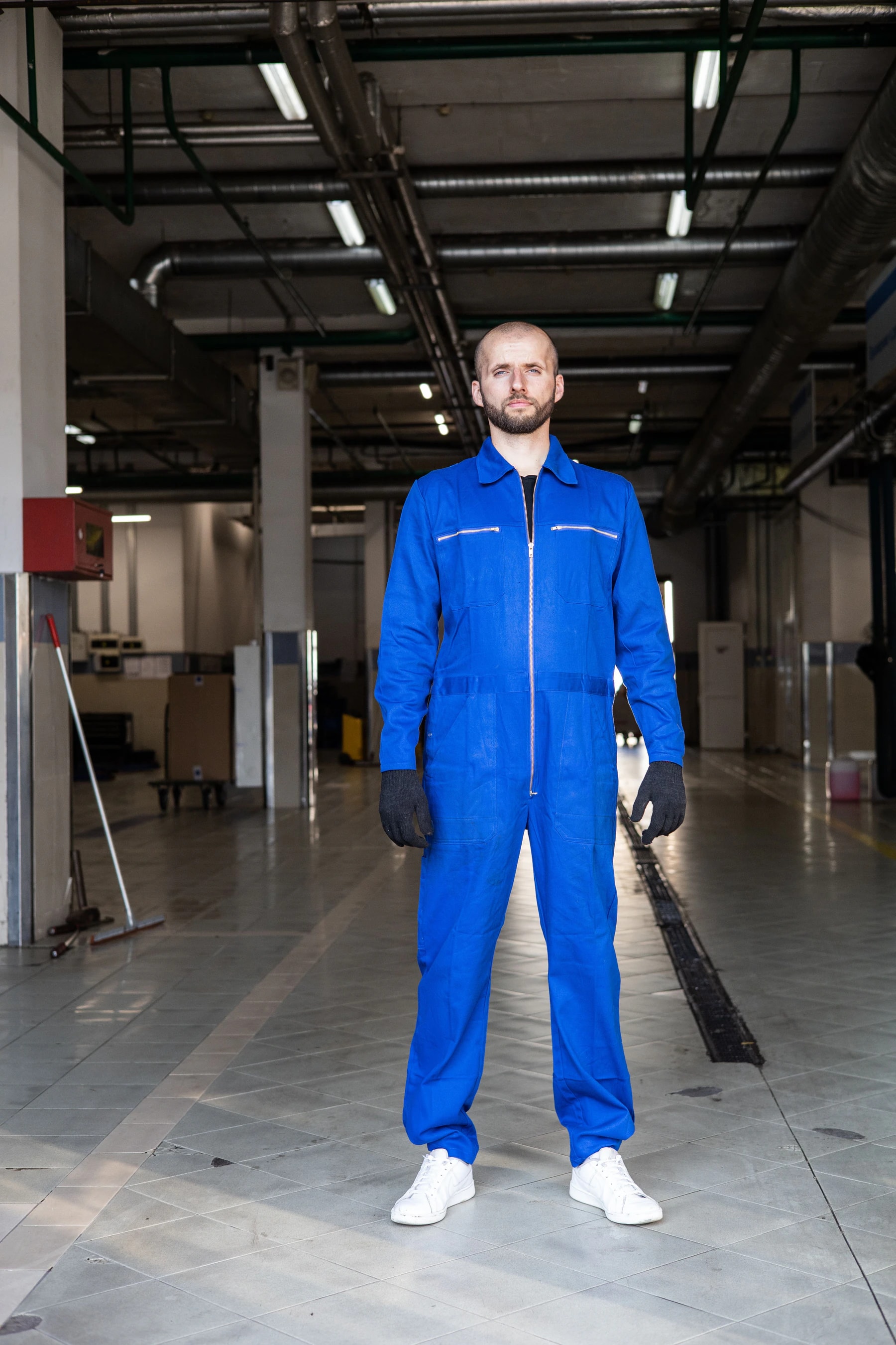Man wearing a one-piece royal blue zip-front work jumpsuit standing in a bright industrial workshop
