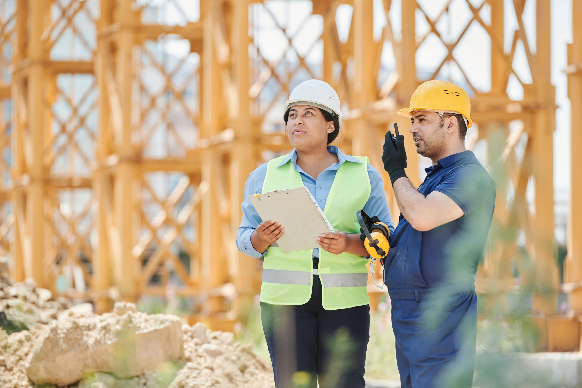 Woman in navy work pants, hi-vis vest and white hard hat holding a clipboard on a construction site